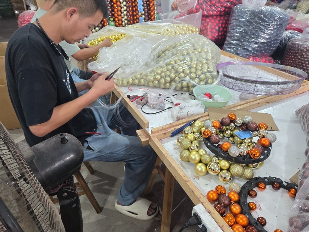 Factory worker hand-assembling custom Christmas ball wreaths with gold and bronze ornaments at a wholesale production bench